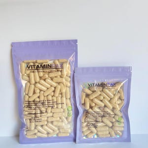 May include: Two sealed bags filled with beige capsules. The bags have a light purple border. The text "VITAMINPAX" is printed on the bags, along with other text describing the product's benefits and ingredients. The capsules are for wellness.