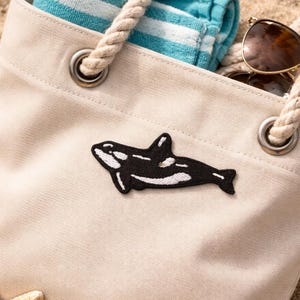 Puede incluir: Un bolso tote de lona beige con un parche de orca en blanco y negro, asas de cuerda y ojales metálicos. La bolsa contiene una toalla a rayas azules y blancas y gafas de sol. Chanclas amarillas y una estrella de mar están en la arena.