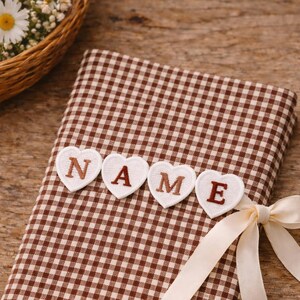 May include: A brown and white gingham fabric-covered book with the word "NAME" spelled out in heart-shaped appliqués. A cream-coloured ribbon is tied around the book, and a basket of flowers is in the background.