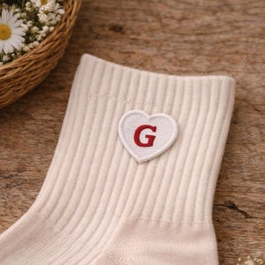 May include: Cream-coloured sock with a ribbed design, featuring a heart-shaped patch with a red "G" initial. The sock is displayed on a wooden surface, with a basket of white flowers in the background. A spool of thread and scissors are also visible.