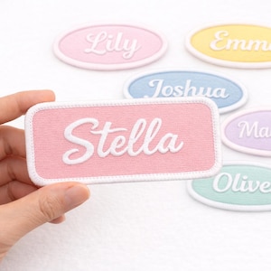 Op de afbeelding: Een hand houdt een roze rechthoekige patch met de naam "Stella" in witte letters. Andere ovale patches met namen als "Lily", "Emma", "Joshua", "Madison" en "Oliver" zijn verspreid, elk in een andere pastelkleur.