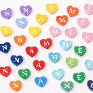 May include: A selection of heart-shaped patches in various colours. Each patch displays a single letter: N, A, M, or E. The patches are in shades such as yellow, orange, red, pink, green, blue, and purple. The letters are in white.
