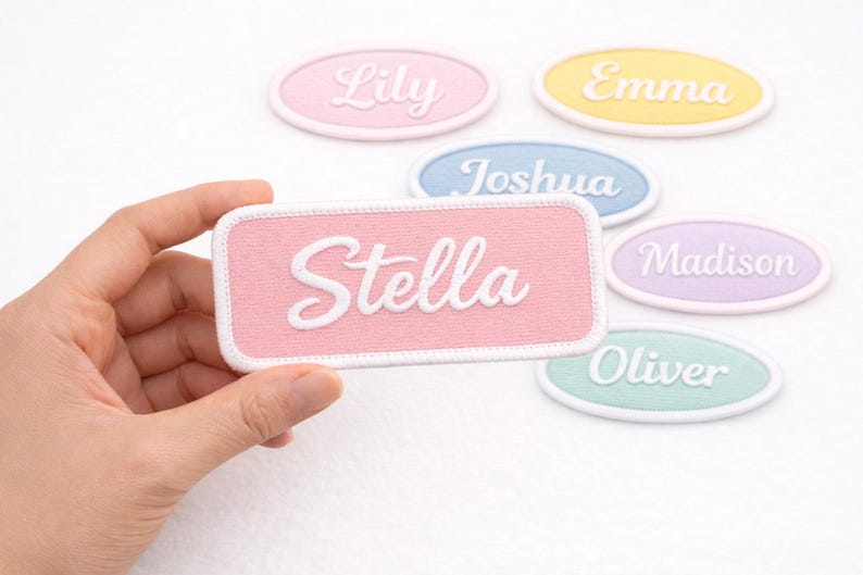 Pu&ograve; includere: Una mano tiene una toppa rettangolare rosa con il nome "Stella" in corsivo bianco. Altre toppe ovali con nomi come "Lily", "Emma", "Joshua", "Madison" e "Oliver" sono sparse, ognuna in un colore pastello diverso.