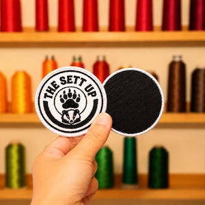May include: Two circular embroidered patches with a white border. One patch features a black bear paw print above a badger face, surrounded by the text THE SETT UP. The second patch is solid black with a white border, showing a textured fabric surface.
