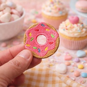 May include: A pink embroidered donut patch with colourful sprinkles and a gold border. Cupcakes, macarons, and marshmallows are in the background. The image has a sweets and treats theme.