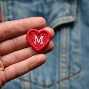 May include: A small, red heart-shaped patch with a white embroidered letter "M" in the centre. The patch is held in a hand, with a denim jacket in the background. The patch is likely an iron-on or sew-on embellishment.