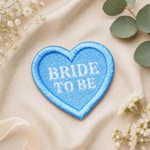 May include: A light blue heart-shaped patch with the embroidered text "BRIDE TO BE" in white. The patch is surrounded by a blue border. The background is a cream-colored fabric, with white flowers and greenery.
