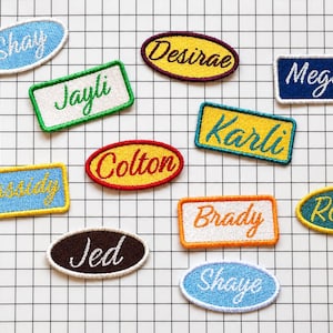 Op de afbeelding: Een verzameling geborduurde naamlabels in verschillende vormen en kleuren. Namen als "Shay", "Jayli" en "Megan" worden in cursief schrift weergegeven. De labels hebben een getextureerd uiterlijk en staan tegen een rasterachtergrond.