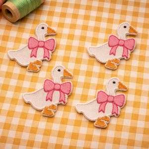 May include: Four white goose patches with pink bows and orange beaks and feet are arranged on a yellow and white checkered surface. Two spools of thread are in the upper left corner. The patches are embroidered.