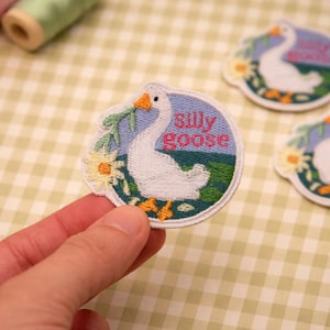 Op de afbeelding: Geborduurde patch met een witte gans, oranje snavel en poten, en bloemendetails, op een blauw en groene achtergrond. De woorden "silly goose" zijn roze geborduurd. De patch is rond.