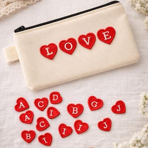 May include: A cream-coloured zipped pouch with the word "LOVE" spelt out in red heart-shaped patches. Additional red heart patches with letters are scattered around the pouch, suggesting customisation options. The background is a white surface.