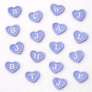 Könnte beinhalten: Eine Sammlung hellblauer herzförmiger Aufnäher, die jeweils mit einem anderen weißen Buchstaben bestickt sind. Die Buchstaben sind B, C, W, J, A, L, H, T und E. Die Aufnäher sind auf einer weißen Oberfläche angeordnet.