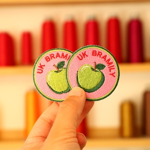 May include: Two circular embroidered patches featuring a light green apple with a brown stem and a dark green leaf on a pink background. The text UK BRAMILY is embroidered in red along the top edge of each patch, which is bordered by a dark green rim.