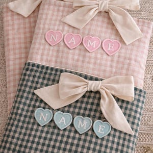 May include: Three gingham fabric items with bows and heart-shaped appliques spelling "NAME". Two are pink, one is green. The bows are a cream colour. The items are on a textured, neutral-coloured surface.