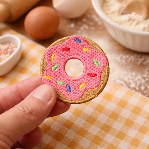 Könnte beinhalten: Ein rosa gestickter Donut-Aufnäher mit bunten Streuseln. Der Aufnäher hat einen braunen Rand und ein Loch in der Mitte. Im Hintergrund sind Backzutaten wie Mehl und Eier.