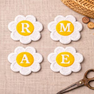 May include: Four white daisy-shaped embroidered patches with yellow centres, each displaying a letter: R, M, A, and E. The patches are arranged on a beige fabric surface, with a pair of scissors and a woven basket in the background.