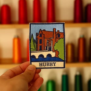 May include: A rectangular embroidered patch featuring a scene of a brown building with a dark roof, a white arched bridge, and green trees against a light blue sky. The word HUBBY is stitched in black capital letters on a cream-coloured banner.