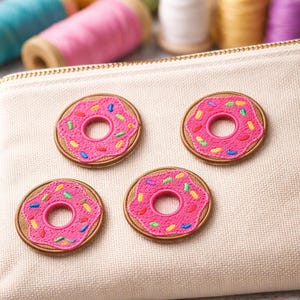 Può includere: Quattro toppe a forma di ciambella rosa con spruzzate colorate sono esposte su una superficie di tessuto beige. Le toppe hanno un bordo marrone e un foro al centro. Forniture per cucito, tra cui rocchetti di filo e forbici, sono sullo sfondo.