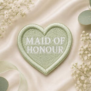 Könnte beinhalten: Ein herzförmiger, salbeigrün gestickter Aufnäher mit dem weißen Schriftzug „MAID OF HONOUR“. Der Aufnäher ist umgeben von weißen Blüten, grünen Eukalyptusblättern und einem hellgrünen Band, alles vor einem cremefarbenen Satinhintergrund.