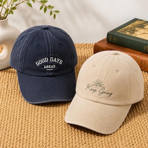 May include: Two baseball caps. One is navy blue with the text "GOOD DAYS AHEAD" in white. The other is beige with the text "Keep Going" and a sun graphic in green. Both caps have a curved peak.
