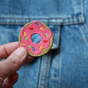 Puede incluir: Parche bordado de donut con glaseado rosa y chispas de colores. El parche tiene un borde marrón y se muestra sobre una chaqueta vaquera azul. Un accesorio divertido y colorido para ropa o bolsos.
