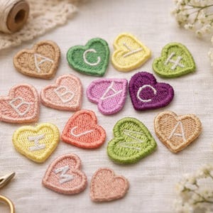 May include: A collection of embroidered heart-shaped patches in various colours, each with a letter of the alphabet. The patches are arranged on a textured, off-white surface, with floral accents and sewing supplies.