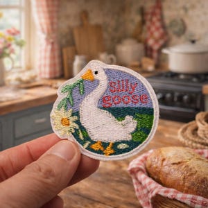 May include: Embroidered patch featuring a white goose with an orange beak and feet, surrounded by a floral design and the words "silly goose." The patch is circular with a blue border and green and yellow accents. The background shows a kitchen setting.