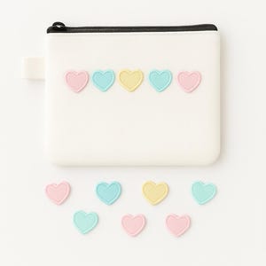 Puede incluir: Una bolsa blanca con cremallera negra y un pequeño lazo de tela. La bolsa está decorada con aplicaciones en forma de corazón de colores pastel en rosa, azul y amarillo. Varias aplicaciones de corazón sueltas están esparcidas debajo.