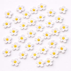 May include: A scattering of small, white daisy flower appliques with yellow centres. The appliques are made of a textured fabric and are arranged on a white background. These floral embellishments are suitable for craft projects.