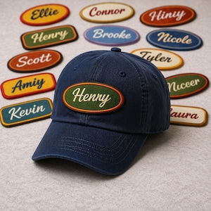 Puede incluir: Una gorra de béisbol azul marino con un parche bordado verde y naranja que dice "Henry". Varios parches con nombres en varios colores y fuentes están esparcidos alrededor de la gorra. Los parches tienen nombres como "Ellie", "Connor" y "Brooke".