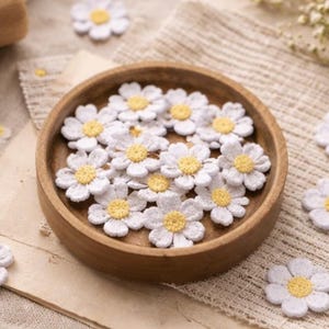 Puede incluir: Aplicaciones de flores de margarita blancas con centros amarillos, presentadas en un cuenco de madera y dispersas sobre una superficie texturizada. Se ve una bobina de hilo beige, lo que sugiere un proyecto de artesanía o bricolaje.