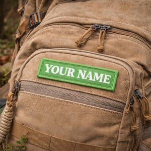 May include: A tan canvas rucksack with multiple pockets, zips, and straps. A green patch with the text "YOUR NAME" in white is sewn onto the front pocket. The rucksack appears to be designed for outdoor activities.