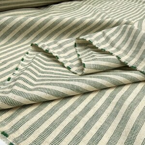 May include: Close-up of a folded fabric with a cream-colored background and green stripes. The stripes are evenly spaced and run diagonally across the fabric. The edges of the fabric are visible, showing a raw, unfinished edge. The fabric appears to be a woven material.