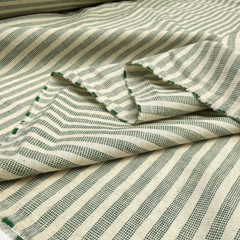 May include: Close-up of a folded fabric with a cream-colored background and green stripes. The stripes are evenly spaced and run diagonally across the fabric. The edges of the fabric are visible, showing a raw, unfinished edge. The fabric appears to be a woven material.