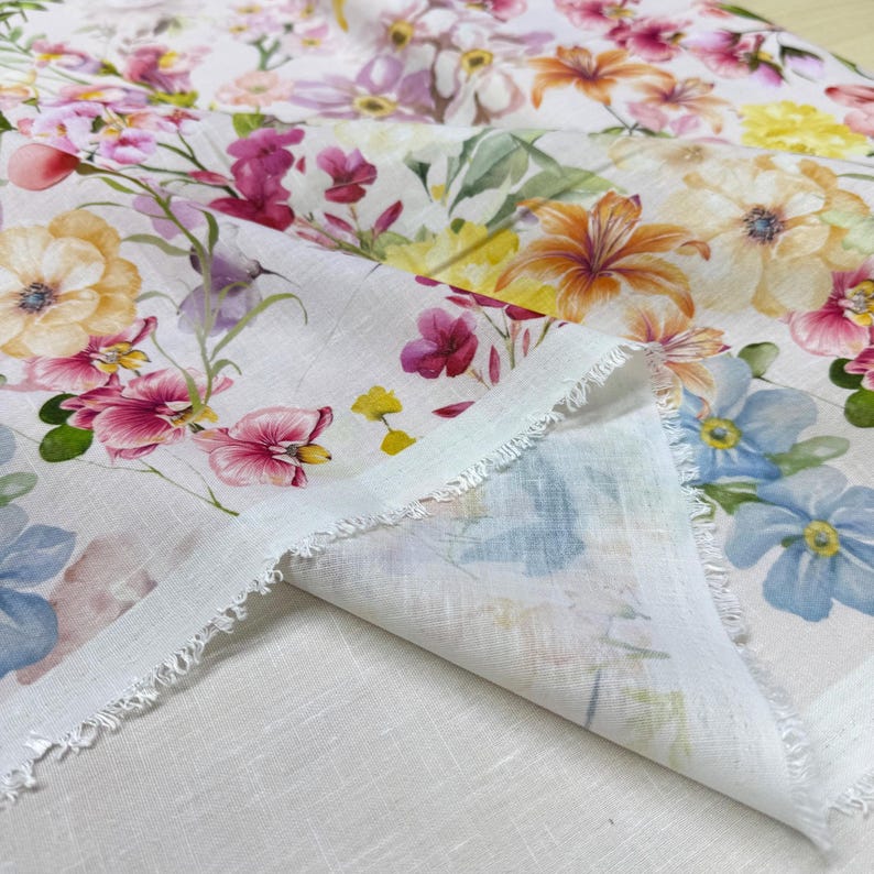 May include: White floral fabric with a watercolor design. The fabric features a variety of flowers in shades of pink, yellow, orange, and blue. The edges of the fabric are frayed, and the fabric is folded over.