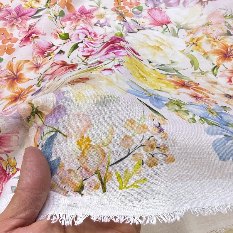 May include: White floral fabric with a vibrant watercolor design. The fabric features a variety of flowers in shades of pink, orange, yellow, and blue. The edge of the fabric has a fringed detail.