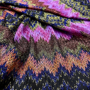 Puede incluir: Primer plano de una tela vibrante con un patrón en zigzag. La tela presenta una mezcla de colores que incluyen negro, azul, naranja, morado y dorado. El material tiene una apariencia brillante y metálica, creando un diseño texturizado y llamativo.