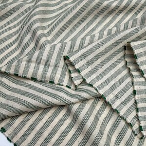 May include: Close-up of a fabric with a cream-colored background and green stripes. The fabric is folded, showing the texture and the green edges. The stripes run diagonally across the fabric.