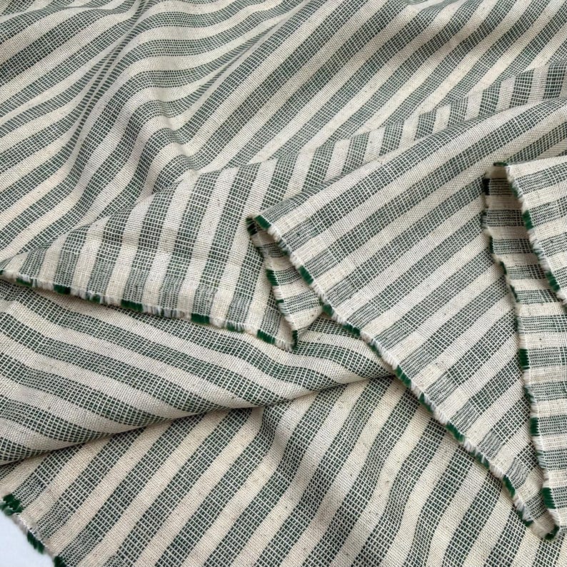 May include: Close-up of a fabric with a cream-colored background and green stripes. The fabric is folded, showing the texture and the green edges. The stripes run diagonally across the fabric.