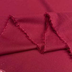 Pode incluir: Close-up de um tecido bordô dobrado com uma textura perfurada. As bordas do tecido estão desfiadas, revelando um aspecto cru e inacabado. O tecido tem um brilho sutil e a cor é um vermelho profundo e rico.