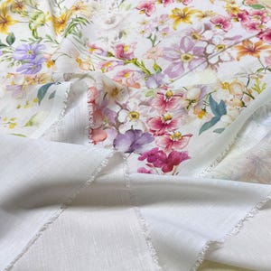 May include: White fabric with a vibrant floral print featuring pink, purple, yellow, and orange flowers. The fabric has a delicate, semi-sheer quality, with a plain white fabric visible underneath. The edges are unfinished, adding a rustic touch.