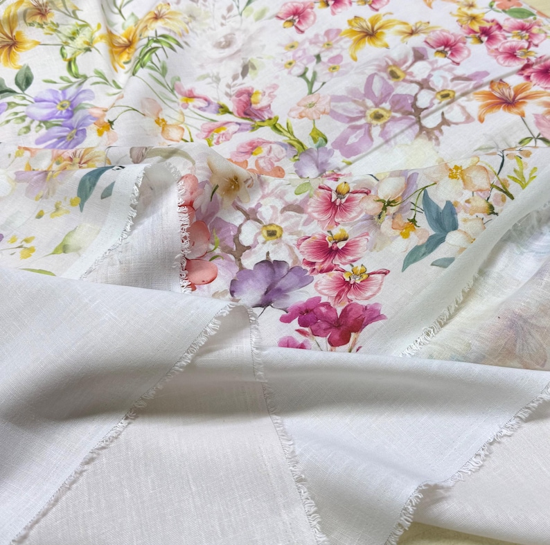 May include: White fabric with a vibrant floral print featuring pink, purple, yellow, and orange flowers. The fabric has a delicate, semi-sheer quality, with a plain white fabric visible underneath. The edges are unfinished, adding a rustic touch.