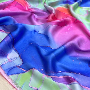 Op de afbeelding: Een levendige stof met een aquareleffect. Het ontwerp toont een mix van kleuren, waaronder tinten blauw, roze, groen en oranje. De stof heeft een zijdeachtige glans, met een vloeiend, abstract patroon. Het algehele effect is artistiek en kleurrijk.