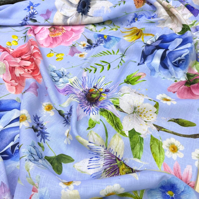 Peut inclure: Un tissu bleu clair orn&eacute; d'un motif floral vibrant. Le design pr&eacute;sente diverses fleurs aux tons rose, blanc, bleu et violet, avec des feuilles vertes. Le tissu est probablement du lin ou une mati&egrave;re l&eacute;g&egrave;re similaire, adapt&eacute; &agrave; l'habillement ou &agrave; la d&eacute;coration int&eacute;rieure.