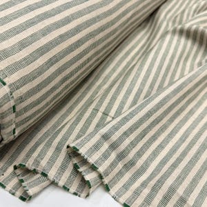 May include: A roll of fabric with a cream-colored background and vertical green stripes. The fabric is folded and rolled, showing the texture and pattern. The edges are finished with green trim. This textile is suitable for various sewing projects.
