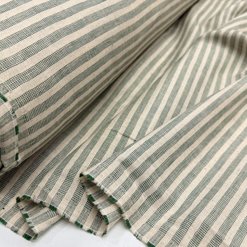 May include: A roll of fabric with a cream-colored background and vertical green stripes. The fabric is folded and rolled, showing the texture and pattern. The edges are finished with green trim. This textile is suitable for various sewing projects.