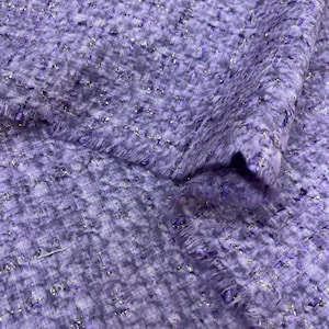 May include: Close-up of a textured fabric in shades of lavender and white, with silver metallic threads woven throughout. The fabric has a soft, slightly fuzzy texture and a frayed edge, suggesting it may be tweed or a similar material.