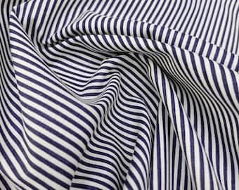 Può includere: Primo piano di un tessuto a righe blu navy e bianche. Il tessuto è drappeggiato, creando pieghe e ombre che aggiungono profondità al design. Le righe sono distanziate uniformemente e corrono verticalmente.