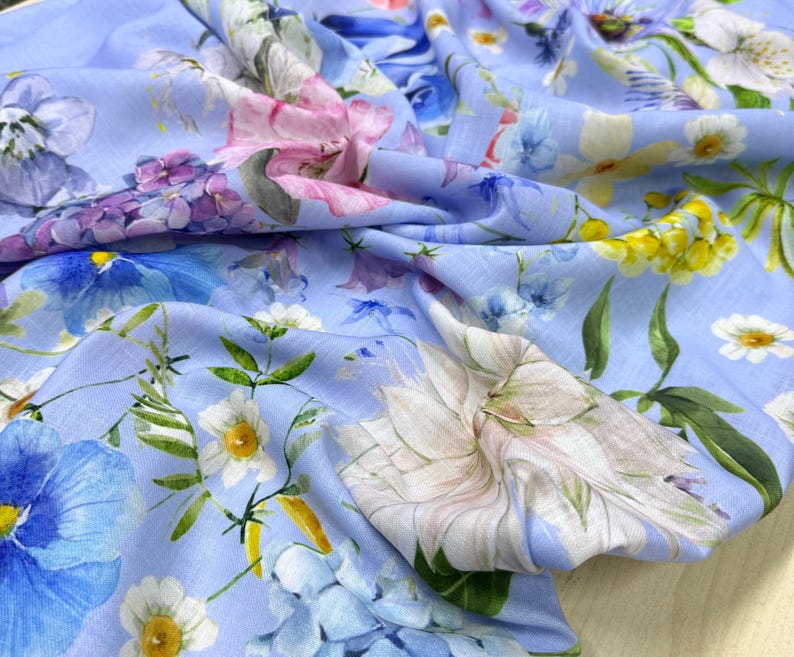 Peut inclure: Un tissu bleu clair avec un motif floral. Le motif pr&eacute;sente une vari&eacute;t&eacute; de fleurs dans les tons de rose, de violet, de blanc et de jaune. Le tissu est drap&eacute;, montrant la texture et le mouvement du mat&eacute;riau.