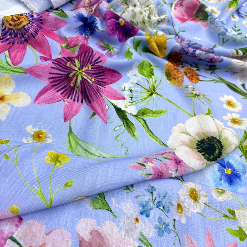Peut inclure: Tissu bleu clair orn&eacute; d'un motif floral color&eacute;. Le design pr&eacute;sente de grandes fleurs de la passion roses, des an&eacute;mones blanches et diverses autres fleurs vives. Des feuilles et des tiges vertes s'entrelacent, cr&eacute;ant une impression botanique anim&eacute;e.